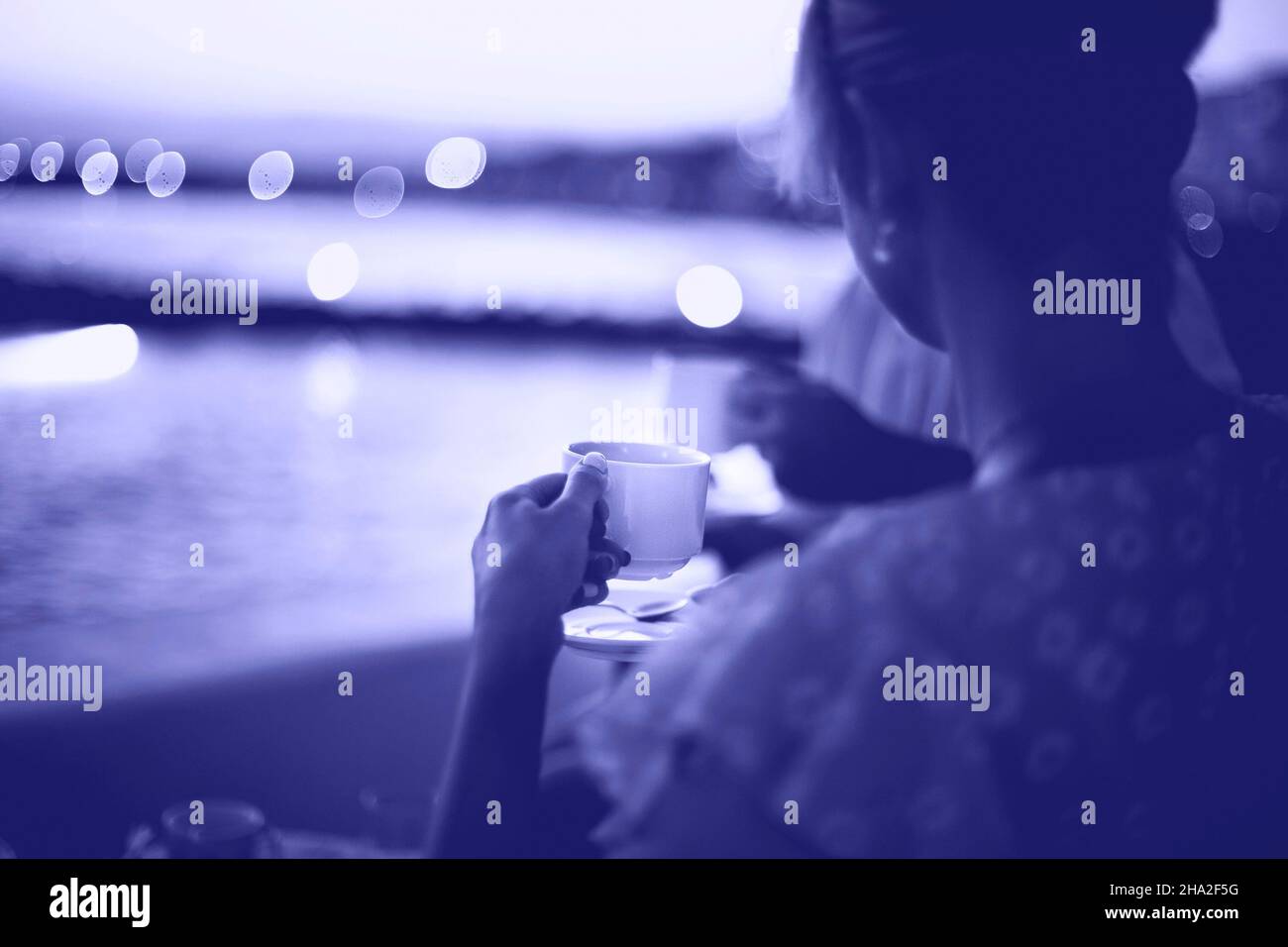 Vue latérale de la jeune femme en robe bleue pour boire du thé tout en étant assise avec un petit ami dans un café extérieur en face de la mer au coucher du soleil.Couleur de 2022 ans, très Banque D'Images