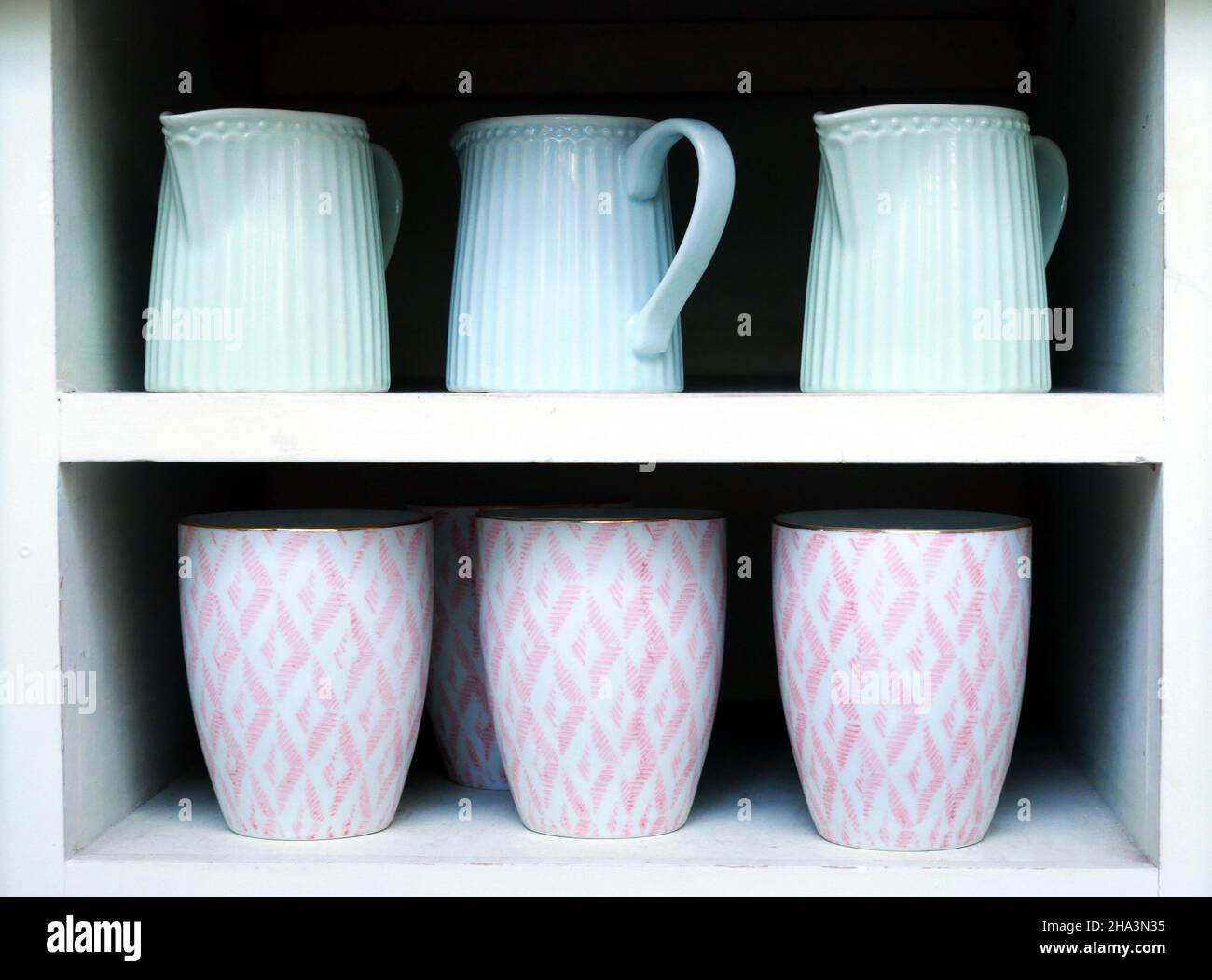 Trois pichets bleu clair sont affichés sur l'étagère supérieure d'une étagère blanche. Trois tasses à motifs roses et blancs sont sur l'étagère du bas. Banque D'Images
