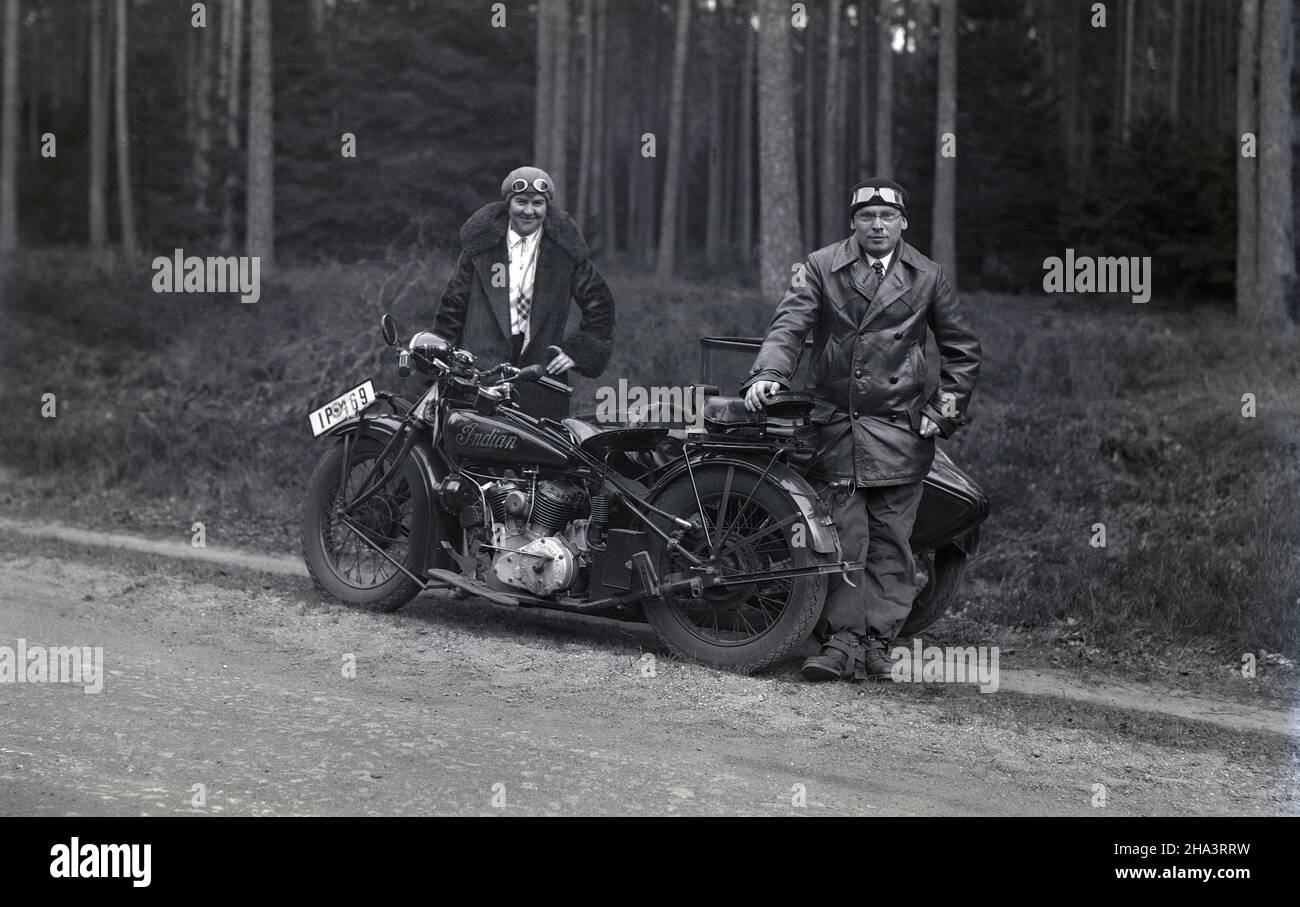 1930s, historique, un gentleman et une dame, portant des manteaux et des  chapeaux avec des gadgets du visage, debout près de leur moto, un scout  indien, avec un side-car, sur une piste