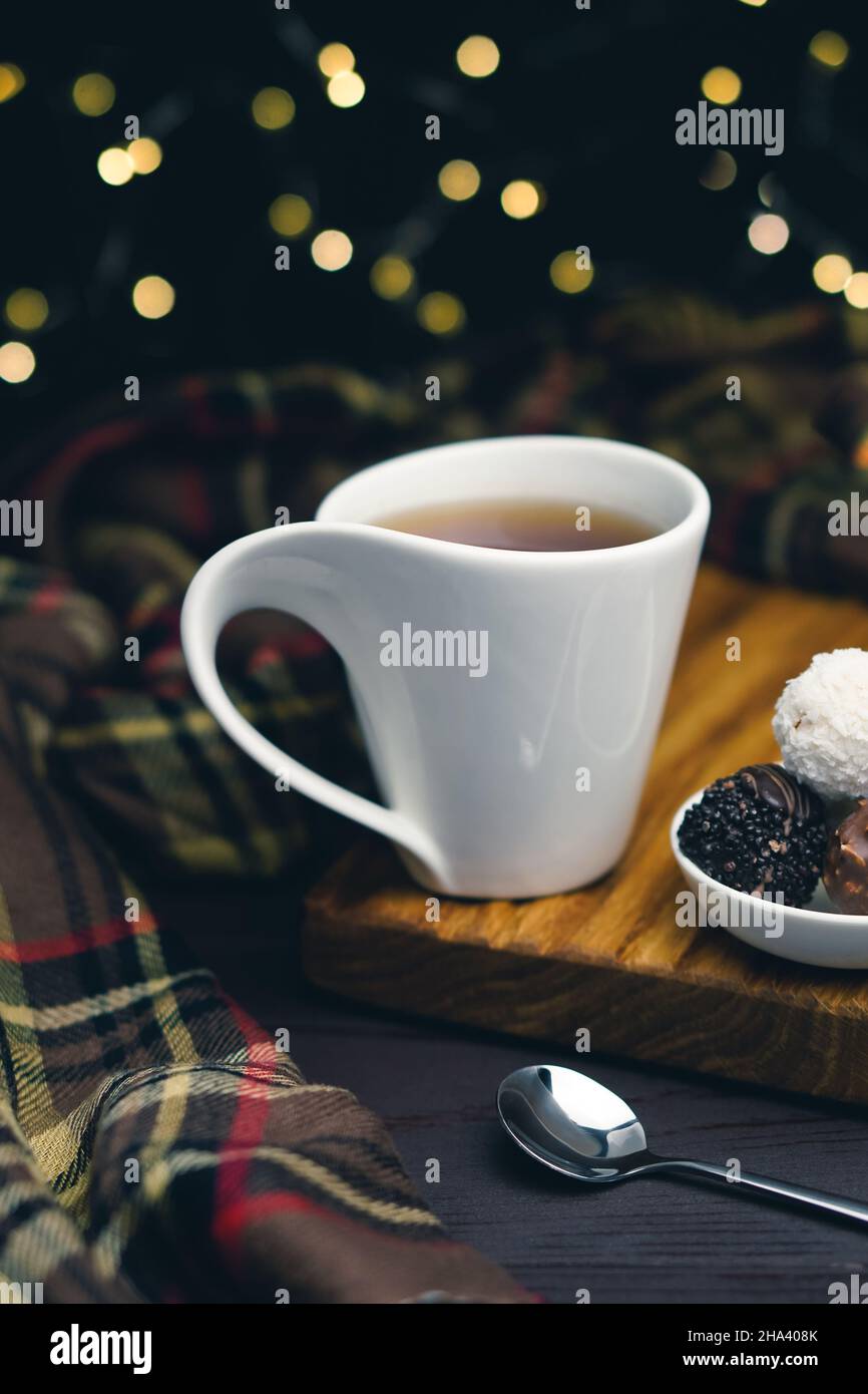 Tasse de thé blanche avec cuillère et bonbons au chocolat sur fond flou avec des lumières de noël Banque D'Images