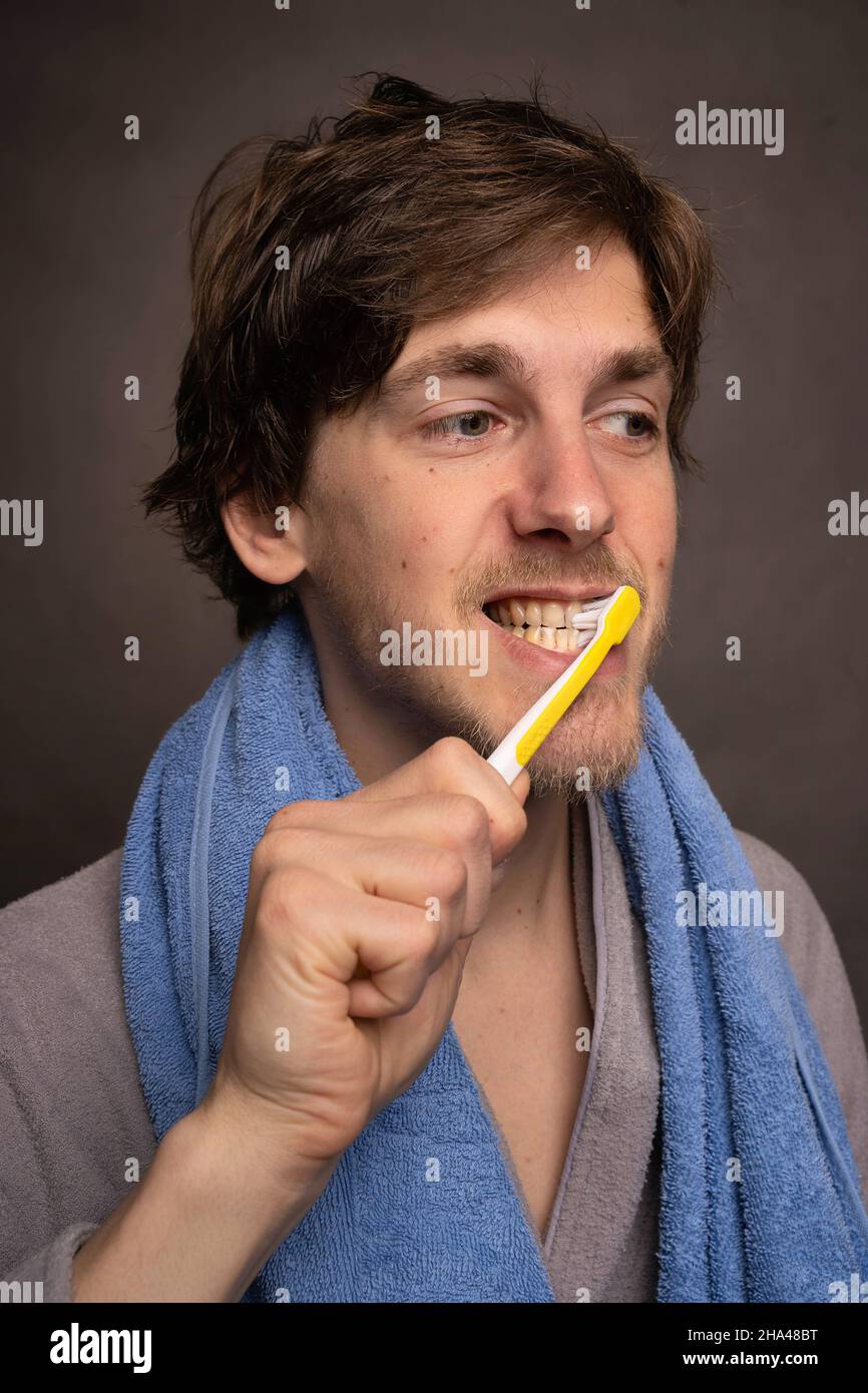 Jeune beau grand homme blanc mince avec des cheveux bruns se brossant ses dents serviette sur les épaules en peignoir gris sur fond gris Banque D'Images