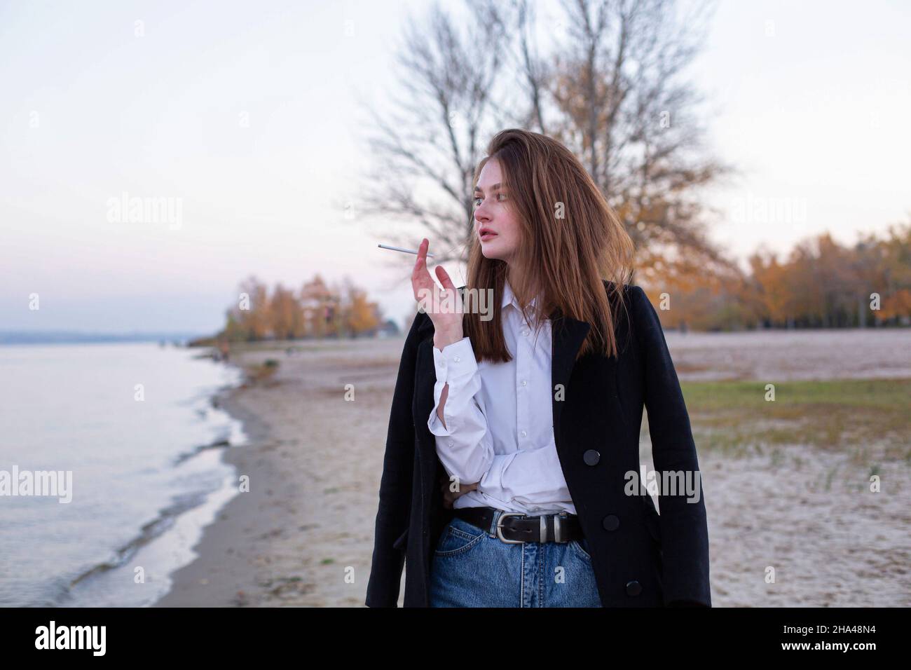 Femme européenne blanche fume, droite et longue, en manteau foncé en automne sur la plage Banque D'Images