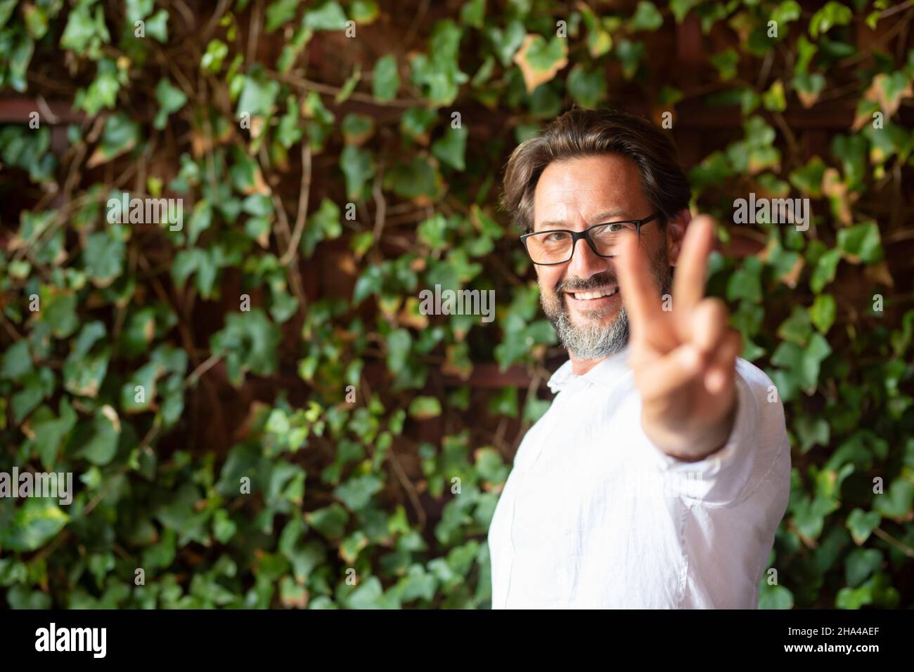 heureux homme mature portant des lunettes et montrant le signe de la victoire des doigts tout en se tenant contre le super-réducteur, homme montrant le signe v. portrait de l'homme souriant montrant le symbole de la paix devant les plantes vertes Banque D'Images
