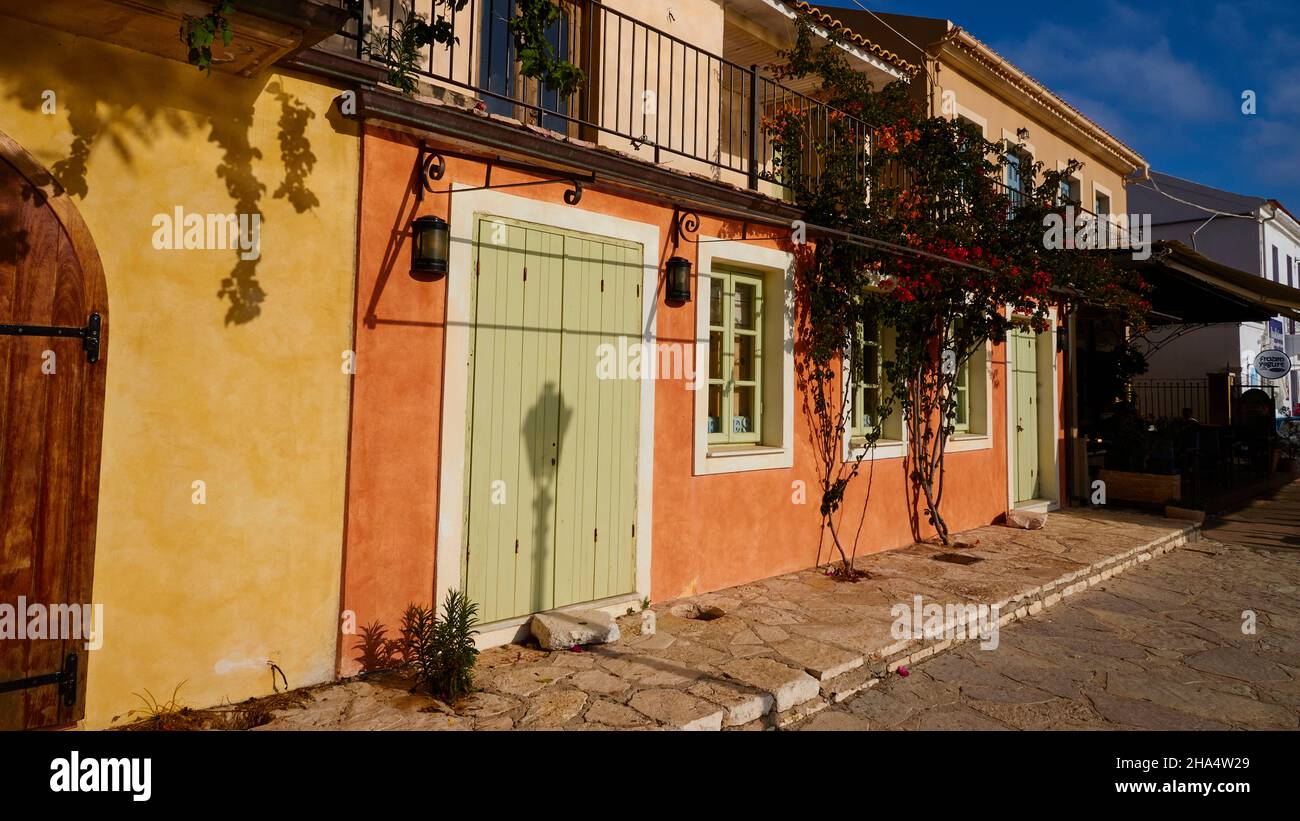 grèce,iles grecques,Iles ioniennes,kefalonia,fiskardo,humeur du matin,ciel partiellement nuageux,rangée de maisons,jaune,vert et ocre dans la lumière du matin,ombre d'une lanterne sur une porte verte,en face de lui un trottoir,en haut à droite petit coin avec un ciel bleu Banque D'Images