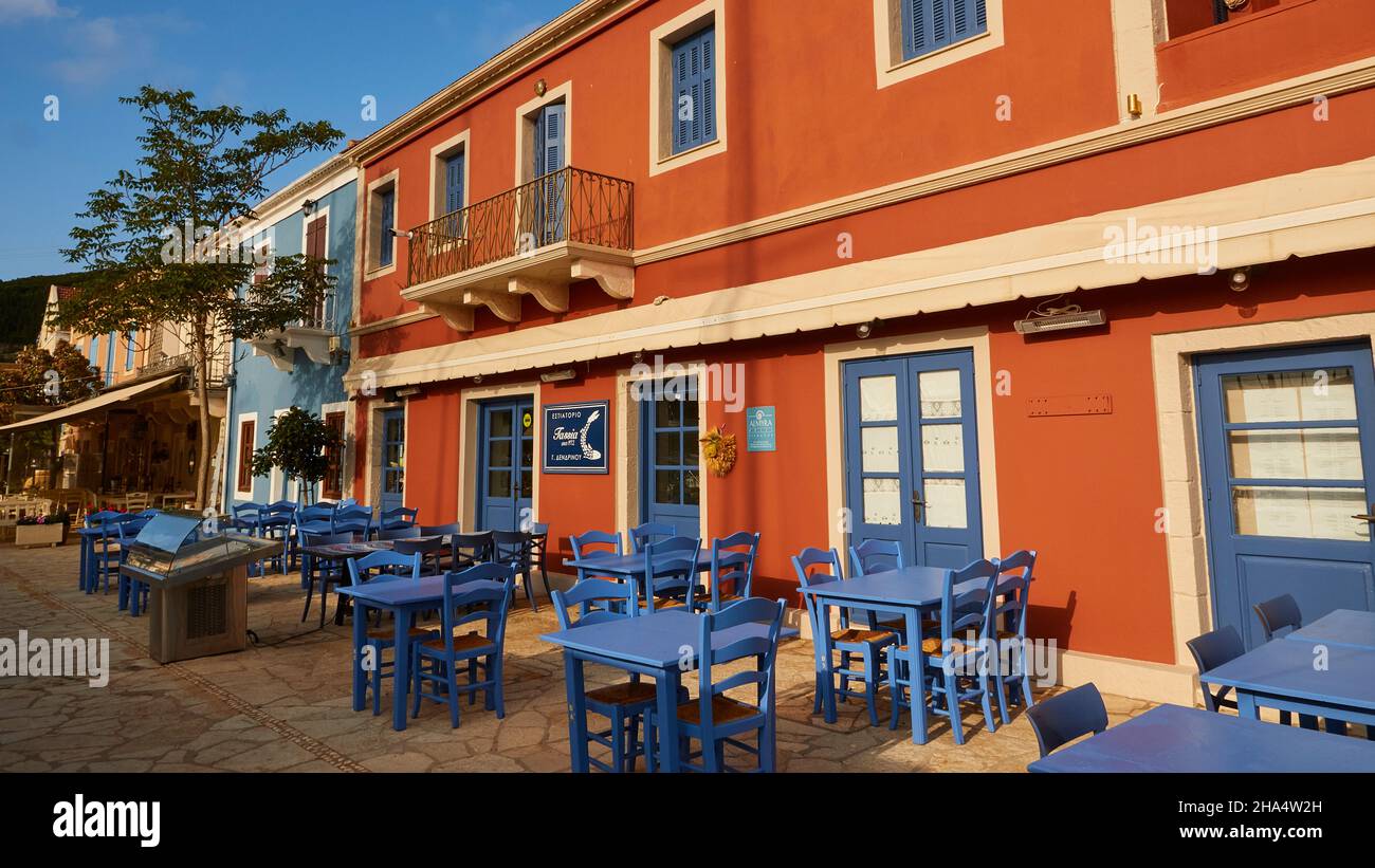 grèce,iles grecques,iles ioniennes,kefalonia,fiskardo,ambiance matinale,ciel partiellement nuageux,café,bar ou restaurant,façade rouge avec lumière matinale devant des tables et chaises bleues vides Banque D'Images