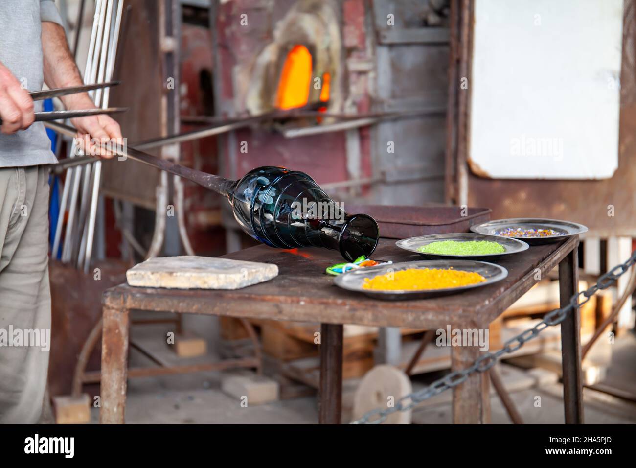 Un artisan vénitien en verre de l'île de Murano travaille sur un vase en verre soufflé sur une table de travail en fer avec une tige en métal. Venise, Italie. Banque D'Images