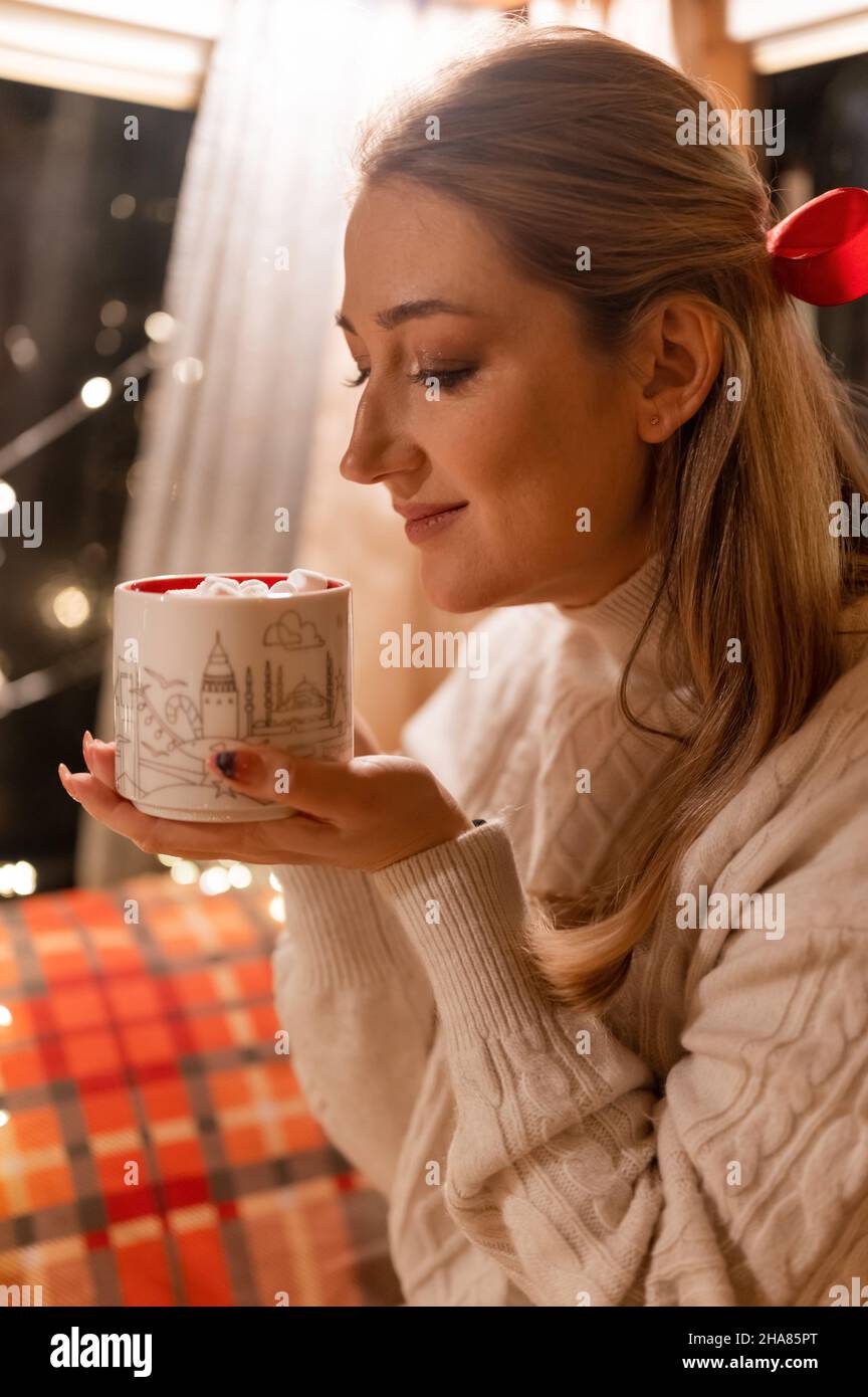 jeune femme souriante et sincère tenant dans les mains et tasse de plaisir de boisson de cacao avec guimauve sur le lit à la maison. concept prenant soin de vous et la rela Banque D'Images