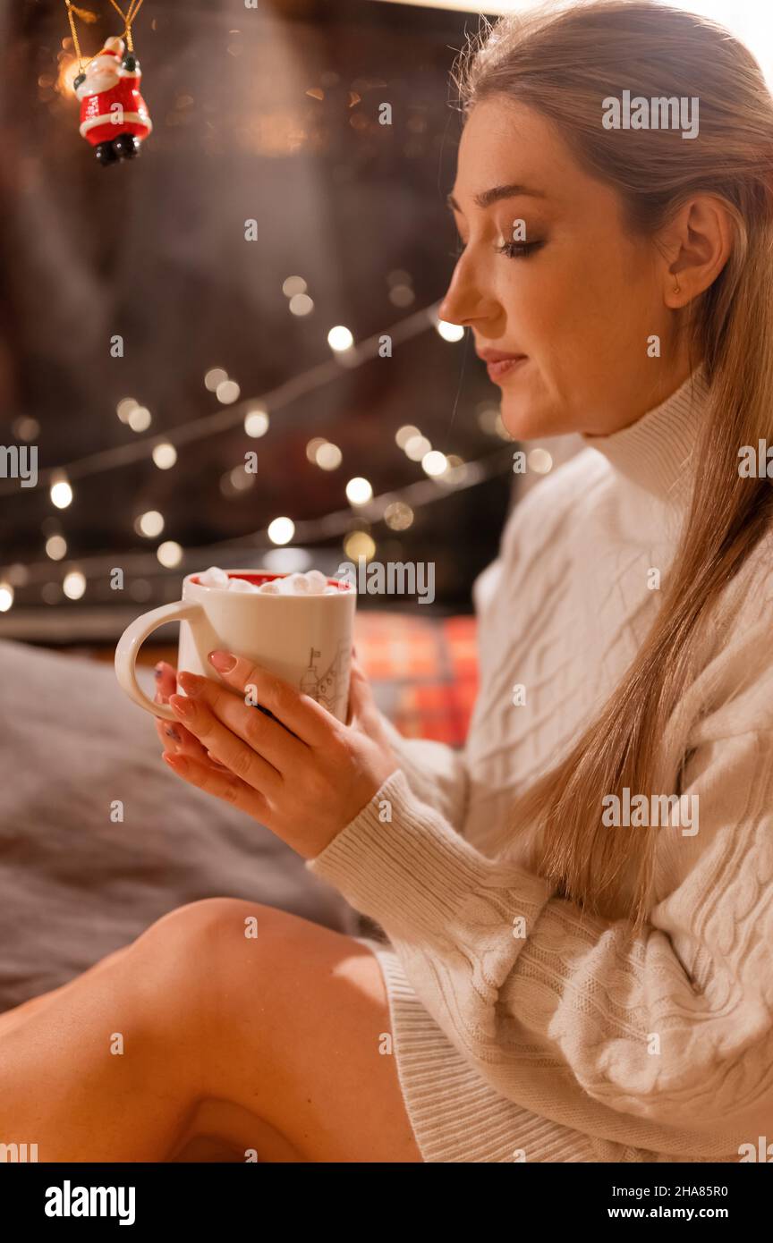 jeune femme souriante et sincère tenant dans les mains et tasse de plaisir de boisson de cacao avec guimauve sur le lit à la maison. concept prenant soin de vous et la rela Banque D'Images