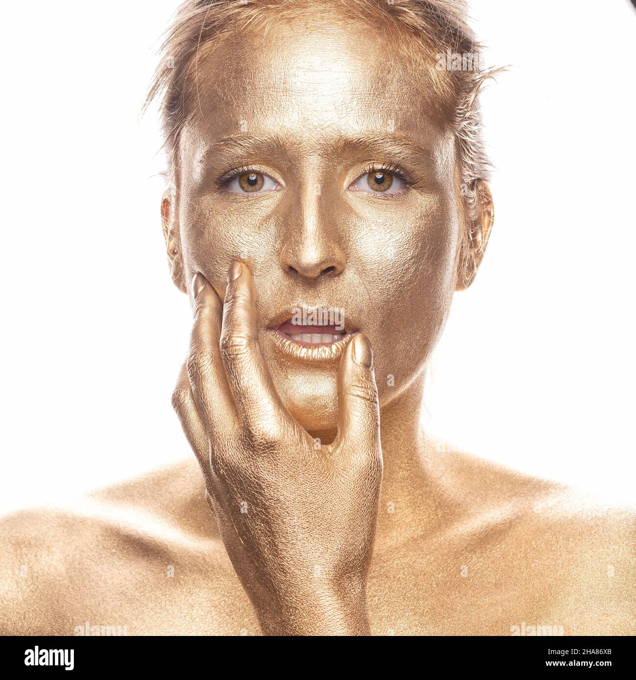 Belle femme peinte or, avec sa main placée sur son visage, près de ses lèvres, regardant dans l'appareil photo Banque D'Images