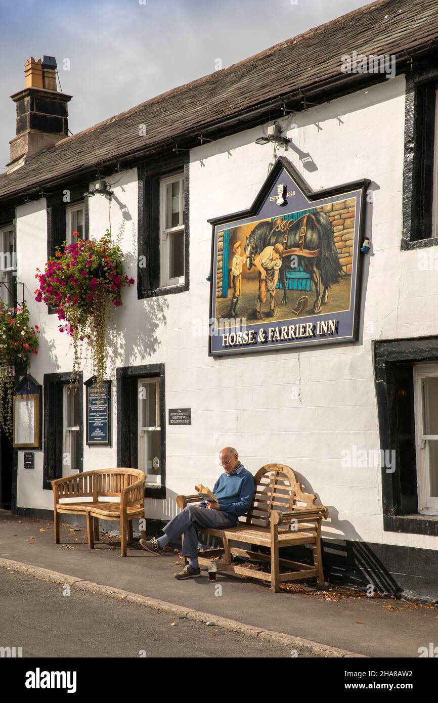 Royaume-Uni, Cumbria, Allerdale, Keswick, Threlkeld,Homme lisant sous le soleil, sous le panneau Jennings Brewery Horse et Farrier pub Banque D'Images