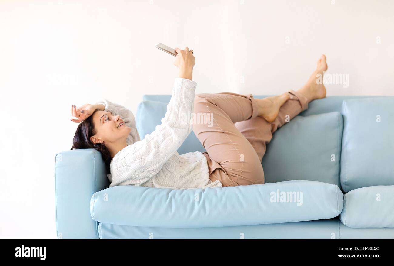 Jeune femme brunette se coucher sur un canapé confortable en position de détente, les jambes lancées, tenant le téléphone mobile, regardant l'écran attentivement, bavardant en ligne dans Banque D'Images