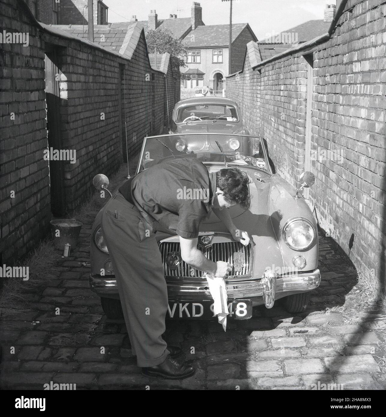 1958, historique, dans une ruelle pavée en brique étroite derrière quelques maisons en terrasse victorienne, un homme nettoyant sa voiture de sport MG à toit ouvert, Angleterre, Royaume-Uni.À côté de la voiture un seau d'eau.À cette époque, MG (garages Morris) était une marque britannique qui produisait des voitures de sport et le MG MGA était une voiture de sport fabriquée de 1955 à 1962.La voiture ici, un MG MGA Twin Cam a été produit de 1958 à 1960. Banque D'Images
