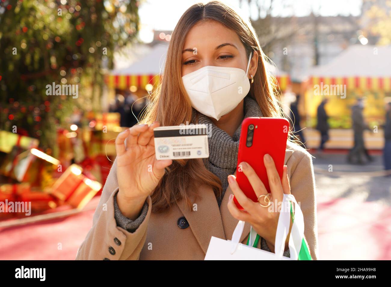 Femme élégante avec KN95 FFP2 masque de protection regardant le numéro de carte de crédit à insérer sur le smartphone pour le paiement en ligne avec les marchés de Noël au dos Banque D'Images