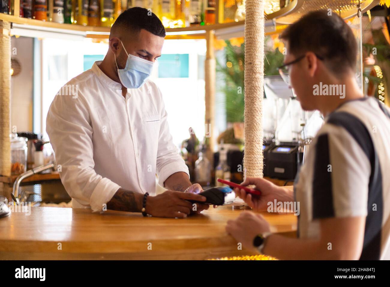 Jeune serveur latino-américain avec masque de visage charge de la barre à un client par paiement sans contact. Banque D'Images