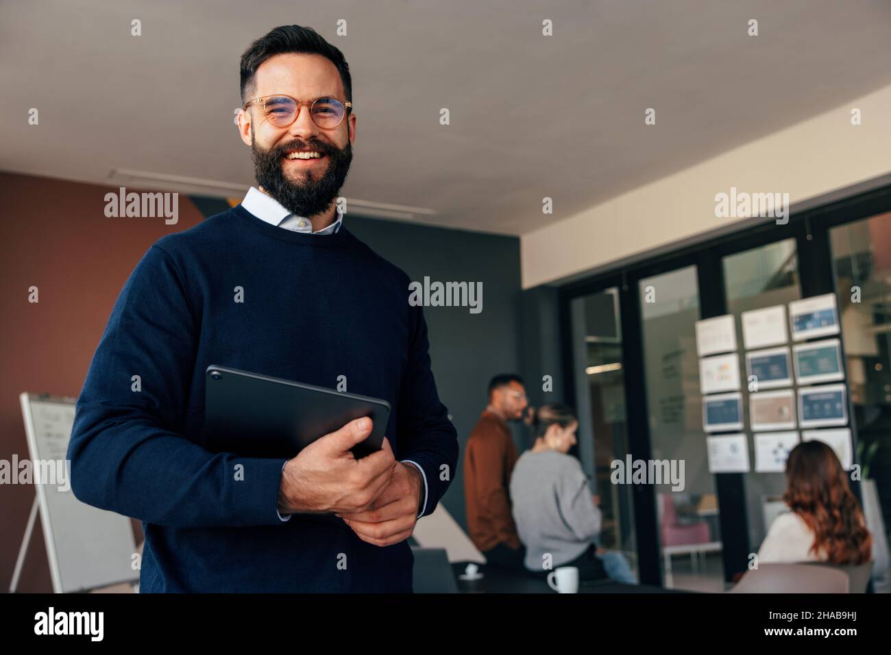 Homme d'affaires souriant à la caméra tout en tenant une tablette numérique.Un jeune homme d'affaires heureux debout dans une salle de réunion moderne avec son collègue Banque D'Images