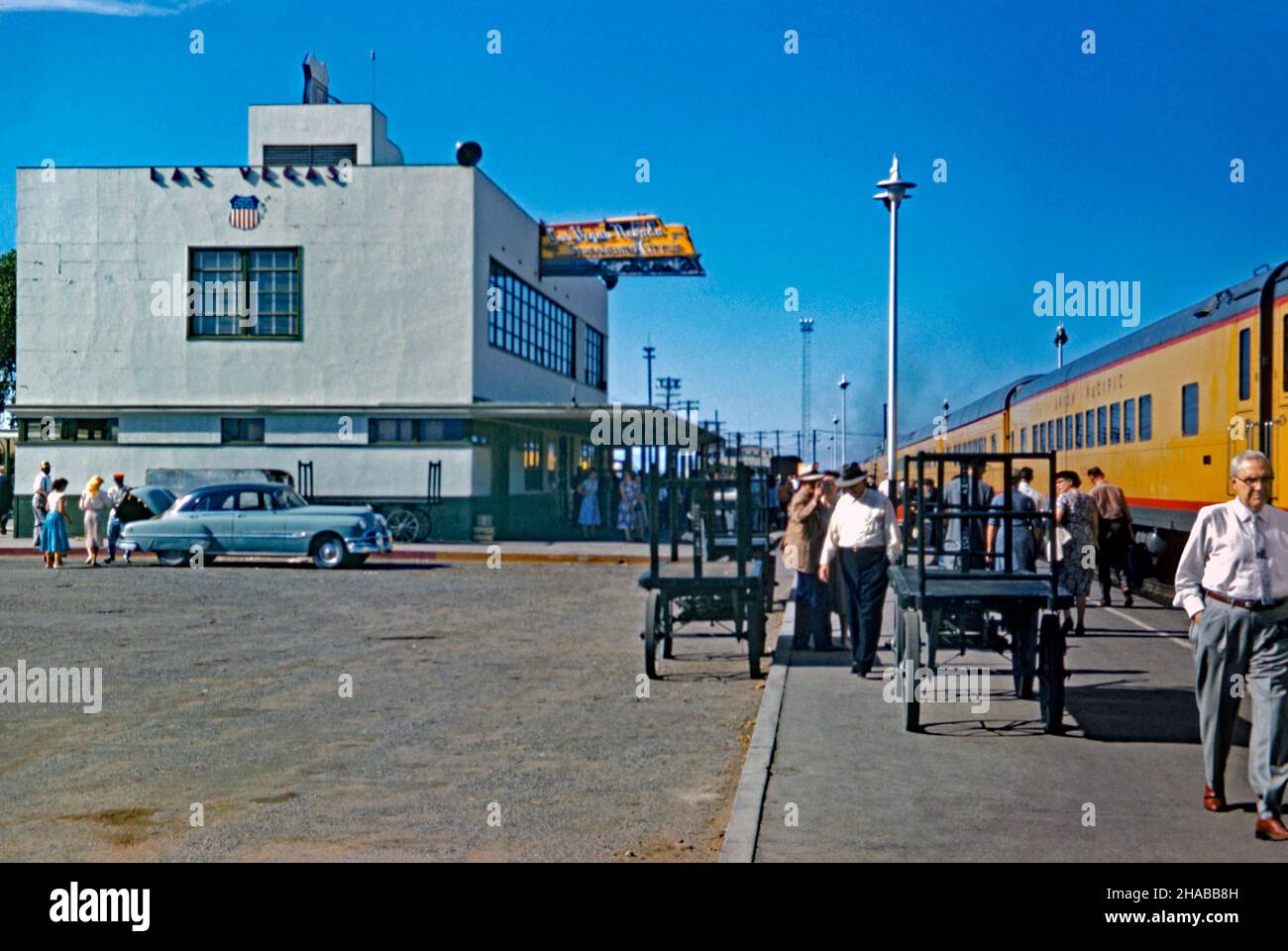 Une scène animée à la gare de Las Vegas, Nevada USA en 1956 – un train Union Pacific est arrivé à la plate-forme.La gare ferroviaire de style «Treamline moderne» a été construite en 1940 et améliorée avec des lumières au néon en 1946 (notez le signe distinctif au néon en forme de moteur de chemin de fer jaune sur le bâtiment).La gare desservait plusieurs trains Union Pacific par jour, reliant Los Angeles à l'ouest et Salt Lake City, puis Chicago et St. Louis à l'est.Cette station a été démolie en 1971 – une photographie vintage de 1950s. Banque D'Images