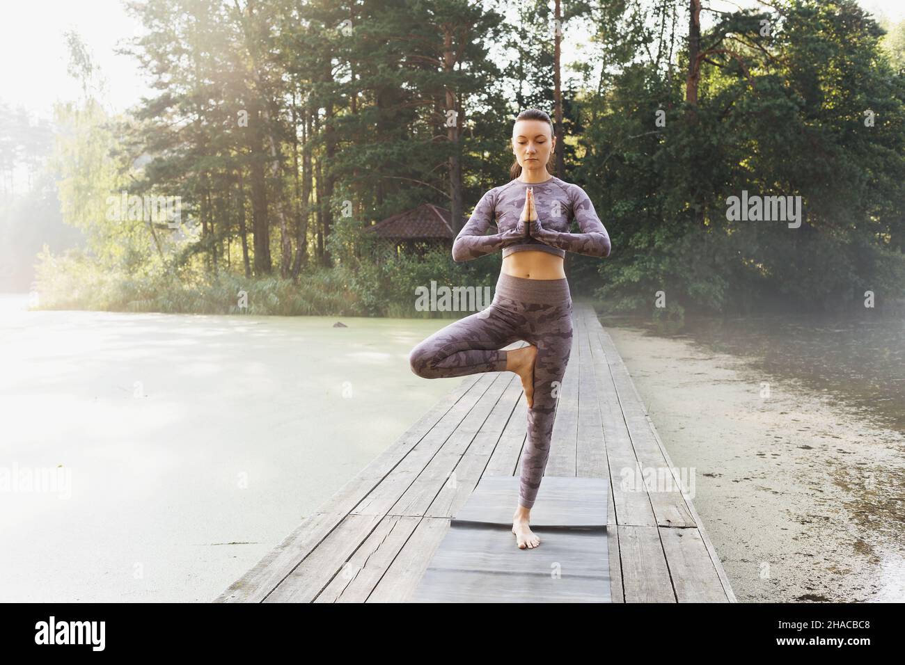 Femme effectue un exercice vrikshasana, pose d'arbre, se tient sur un tapis sur un pont en bois, un matin d'été dans le parc Banque D'Images