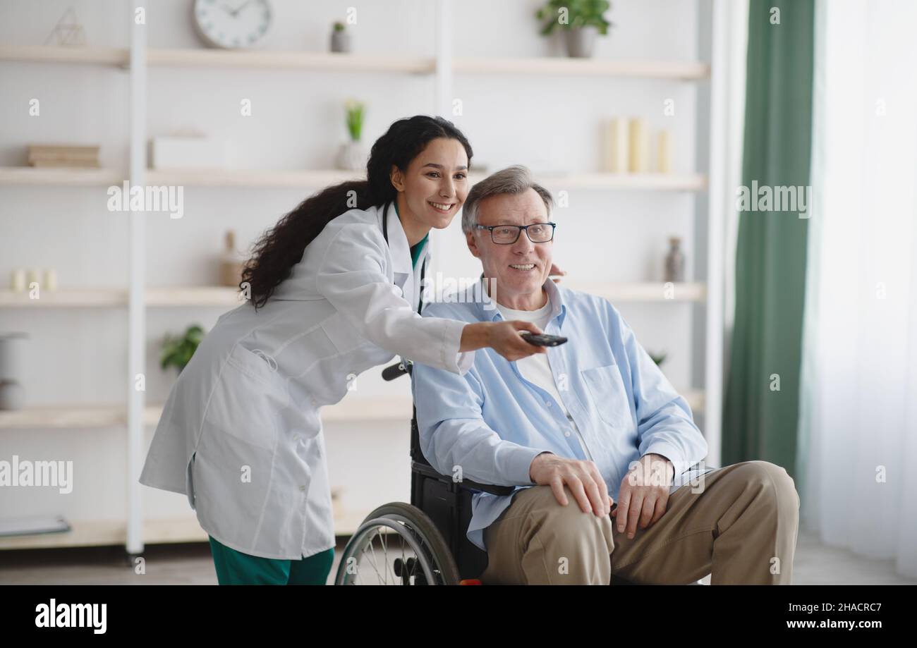 Jeune infirmière avec télécommande et commutation de chaînes à la télévision pour homme âgé en fauteuil roulant à la maison de retraite Banque D'Images