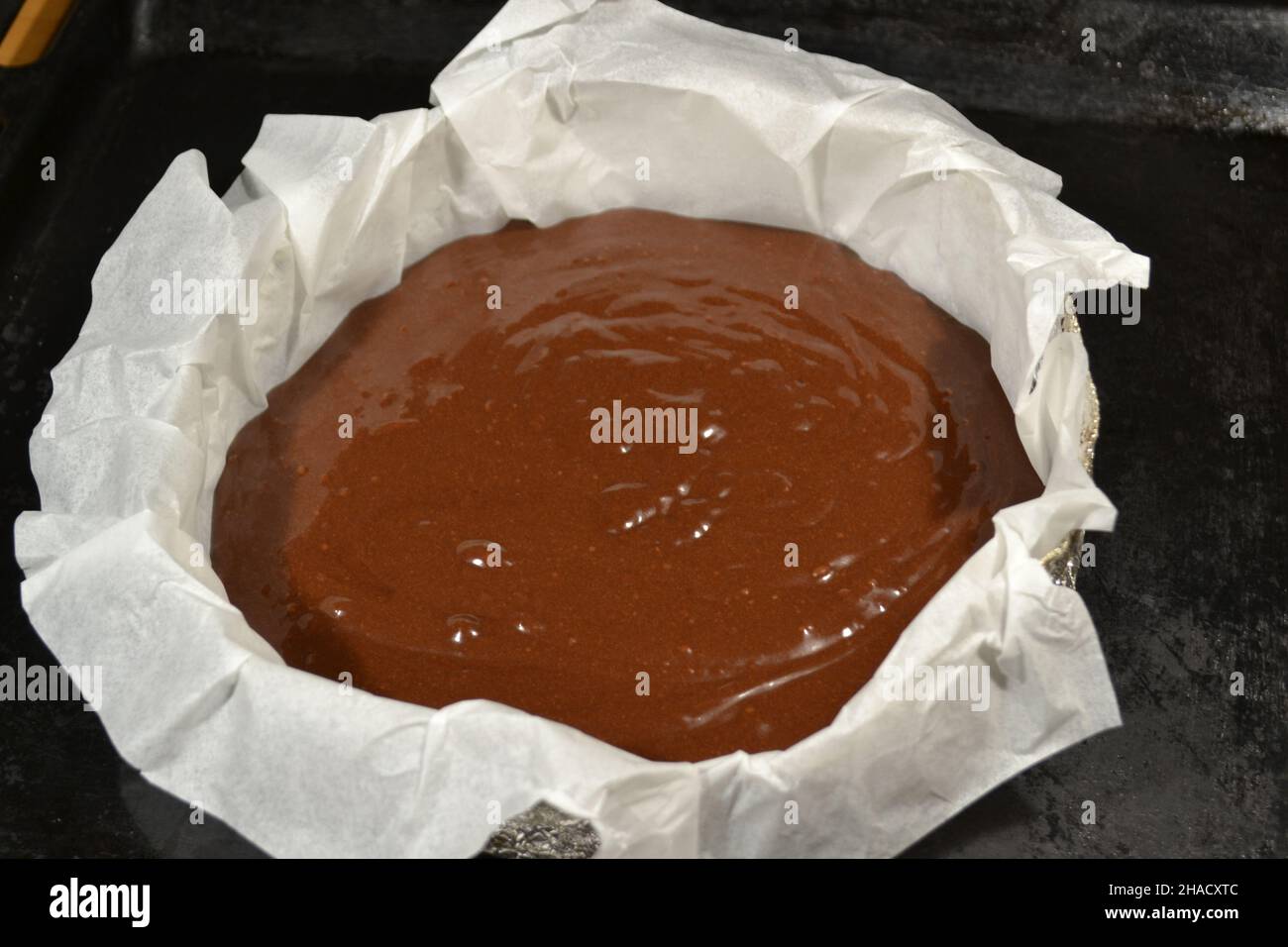 Pâte au chocolat dans un moule de cuisson en aluminium rond fait maison recouvert de papier blanc pour cuire une tortilla à gâteau sur fond noir. Banque D'Images
