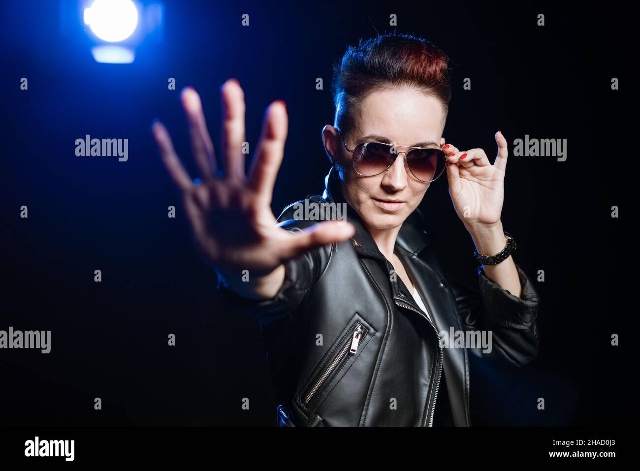 Arrêtez, je ne veux pas voir.Portrait bloquant femme avec cheveux courts et maquillage en cuir noir veste de style décontracté, debout avec un geste d'arrêt dans les lunettes de soleil.Arrière-plan noir avec lumière bleue Banque D'Images