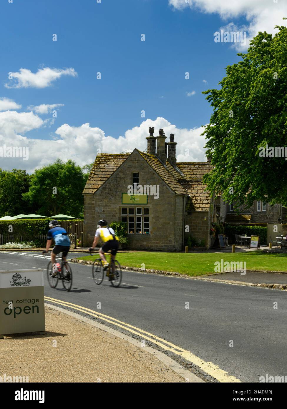 2 cyclistes sur la route de campagne passant par le beau café de salon de thé de chalet dans le pittoresque village rural ensoleillé - B6160, Bolton Abbey, Yorkshire Dales, Angleterre, Royaume-Uni. Banque D'Images