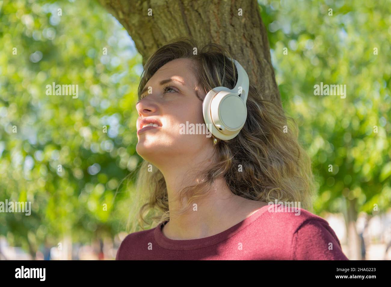 Une femme écoute de la musique à l'aide d'un casque sans fil tout en s'appuyant sur un arbre dans un parc Banque D'Images