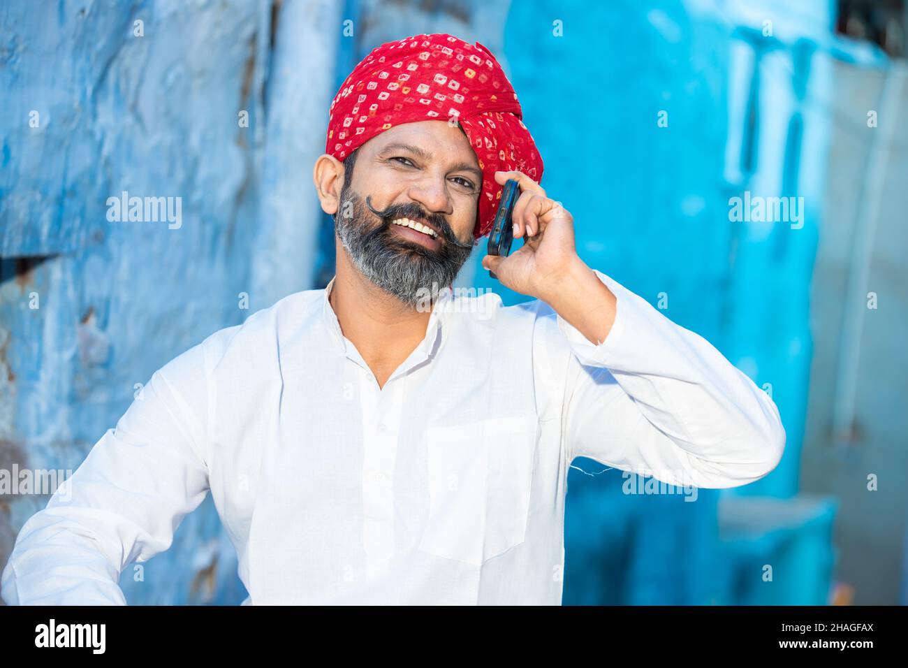 Un homme adulte du nord indien heureux avec barbe et turban rouge parlant sur smartphone, souriant des hommes traditionnels appelant sur smartphone android pendant que s. Banque D'Images