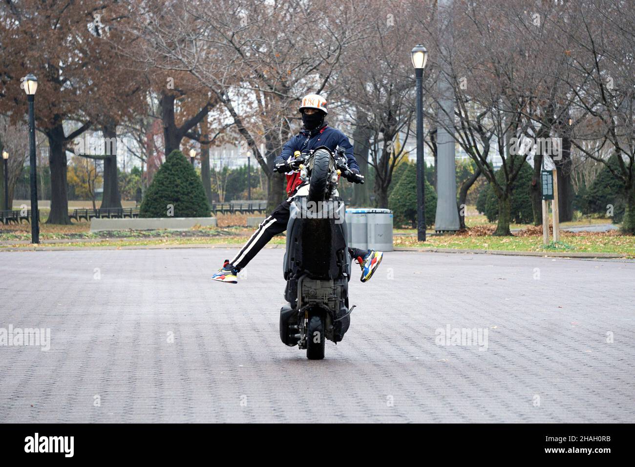 Un motard adolescent avec un casque et un masque fait une roue sur son Yamaha Tmax pendant que ses jambes sont hors de la moto.Dans un parc à Queens, New York. Banque D'Images