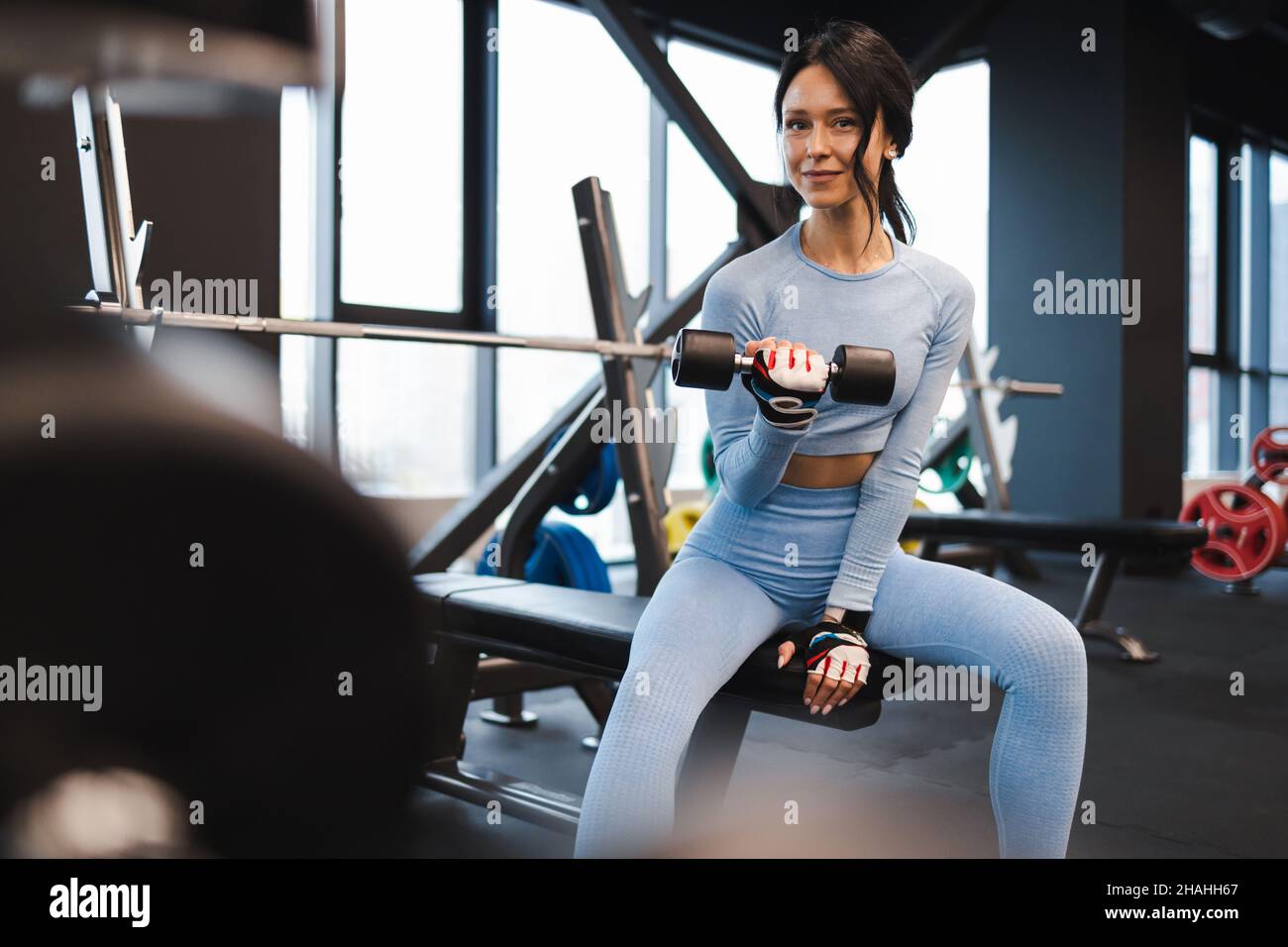 Une femme secoue des biceps levant une haltère tout en étant assise sur un banc dans la salle de gym Banque D'Images