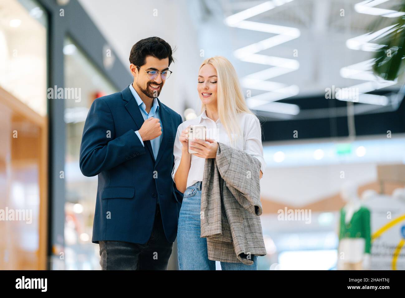 Heureux beau jeune couple en utilisant le téléphone mobile debout dans le hall du centre commercial.Homme barbu en lunettes et femme blonde achetant au centre du magasin. Banque D'Images