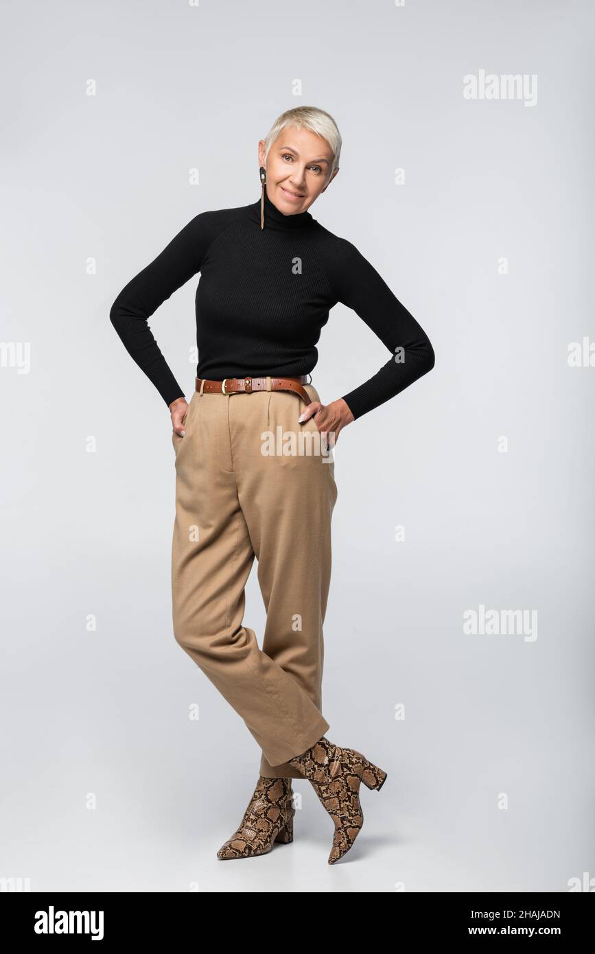 pleine longueur de femme âgée souriante en pantalon beige, col roulé noir  et bottes à imprimé animal posant avec les mains dans des poches sur le  gris Photo Stock - Alamy