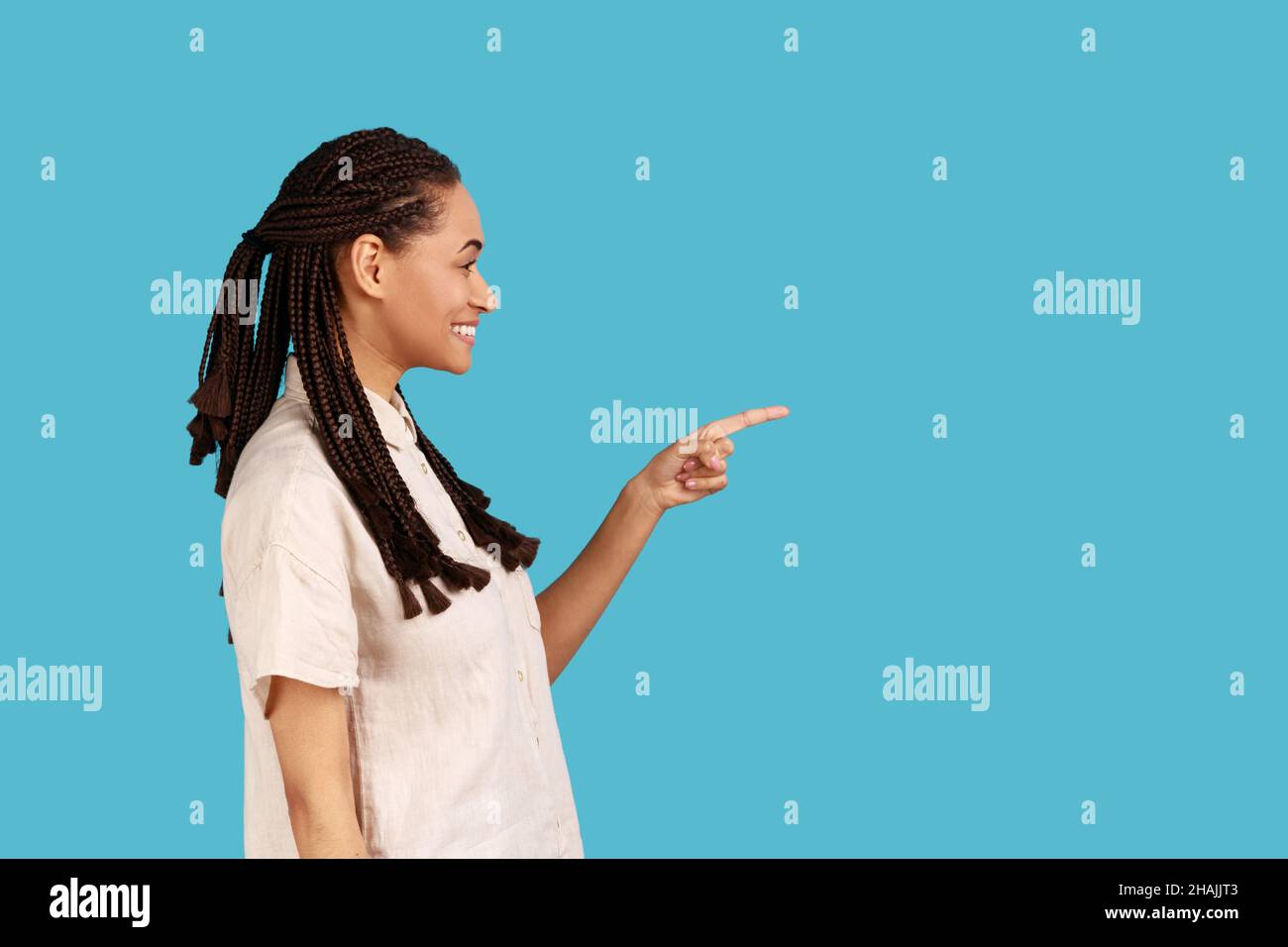 Vue latérale d'une femme souriante avec des dreadlocks noirs pointant vers l'extérieur avec le doigt, montrant un espace vide pour votre publicité, portant une chemise blanche.Studio d'intérieur isolé sur fond bleu. Banque D'Images