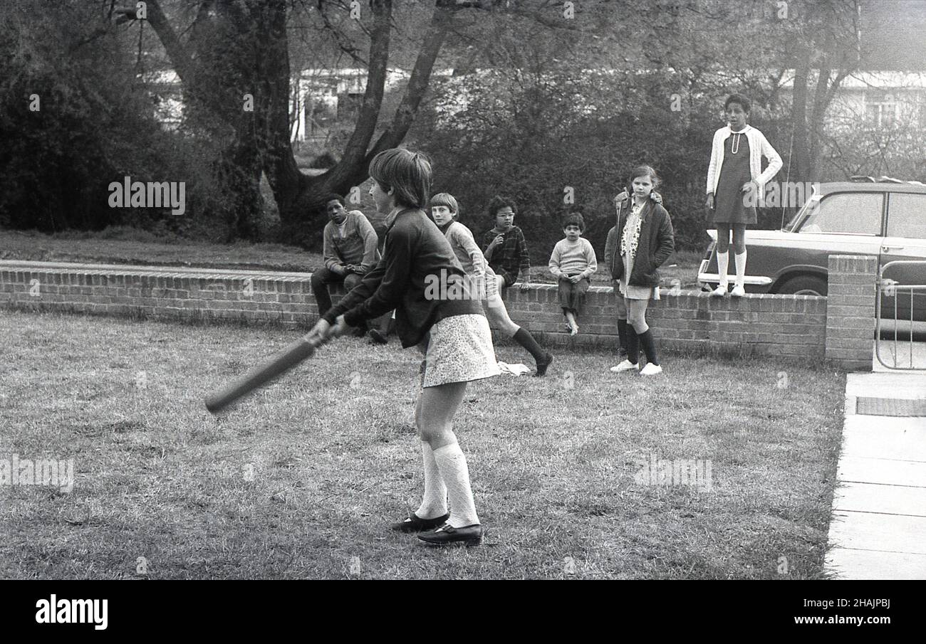 Fin 1960s, historique, dans une banlieue de Londres, des enfants jouant sur un bout d'herbe à l'extérieur de la rue, une jeune fille balançant une batte de cricket, avec d'autres enfants assis sur un mur bas d'observation, Angleterre, Royaume-Uni. Banque D'Images