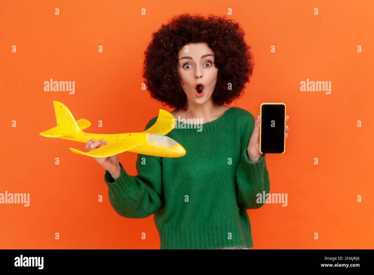 Femme choquée avec la coiffure d'Afro portant un chandail vert décontracté debout tenant l'avion jaune et le téléphone cellulaire avec l'affichage vide, étant profondément étonné.Studio d'intérieur isolé sur fond orange. Banque D'Images