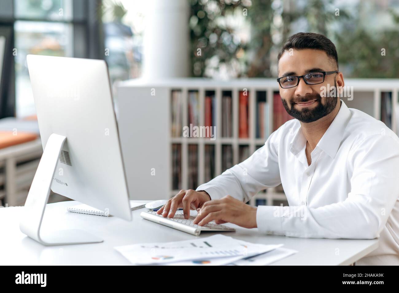 Homme d'affaires indien élégant et réussi, directeur, employé de bureau avec barbe et lunettes.Propriétaire d'affaires assis à une table dans un bureau moderne, en utilisant un ordinateur, regardant l'appareil photo, souriant Banque D'Images
