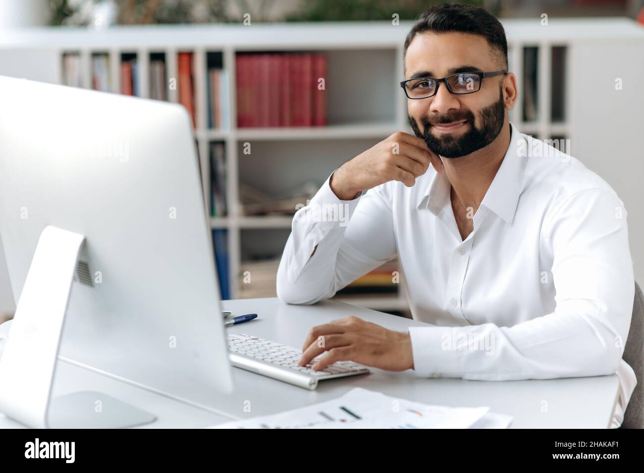 Portrait réussi intelligent heureux beau homme d'affaires indien, indépendant ou directeur avec barbe, portant une chemise blanche, assis à table avec ordinateur au bureau, regardant l'appareil photo et souriant amical Banque D'Images