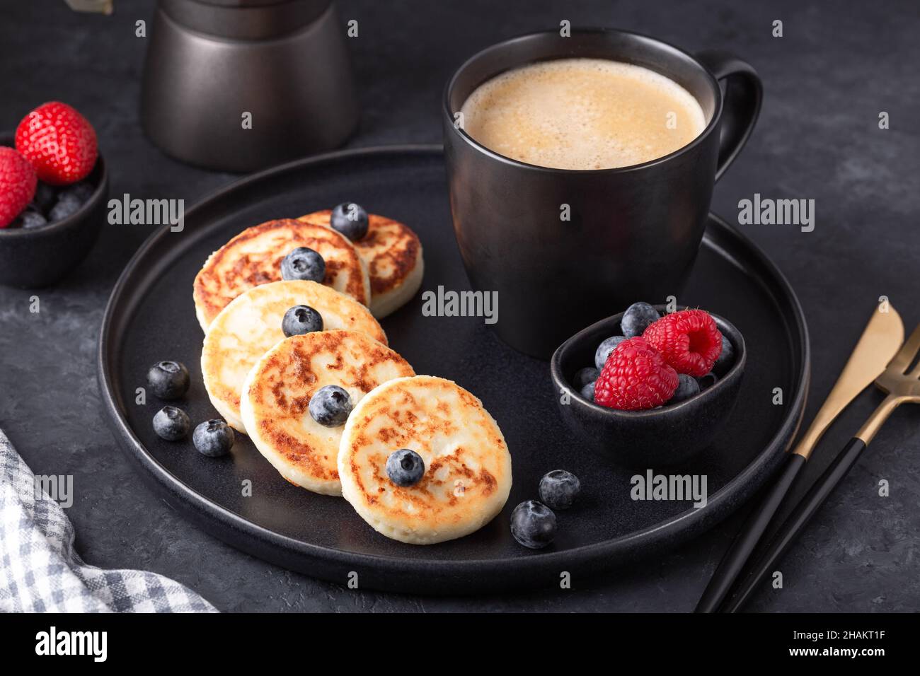Crêpes au fromage cottage avec des framboises fraîches et des bleuets sur une assiette en céramique sombre.Une tasse de café et une cafetière Banque D'Images