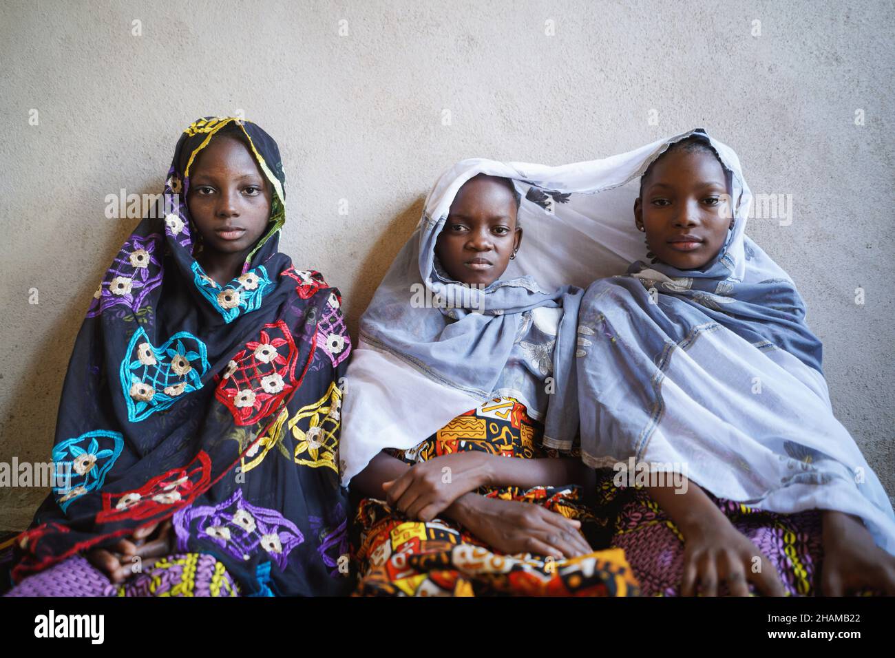 Trois filles africaines s'assoient ensemble et se posent avec des vêtements traditionnels à double paroi sur fond de mur rugueux Banque D'Images