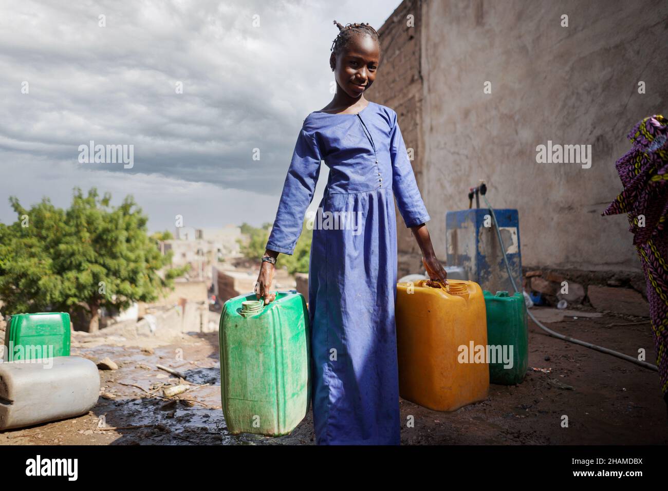Une jeune fille africaine transporte trois conteneurs d'approvisionnement en eau à l'extérieur dans un village africain au Mali Banque D'Images