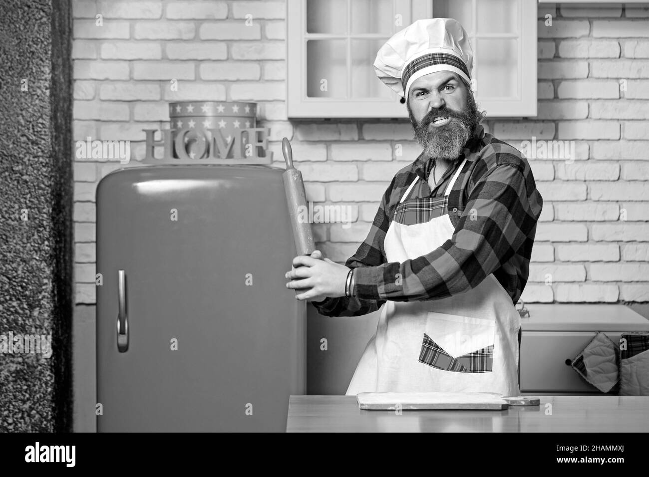 Fou jeune homme chef cuisinier ou boulanger homme en tablier blanc chefs chapeau posant dans le fond de la cuisine. Banque D'Images