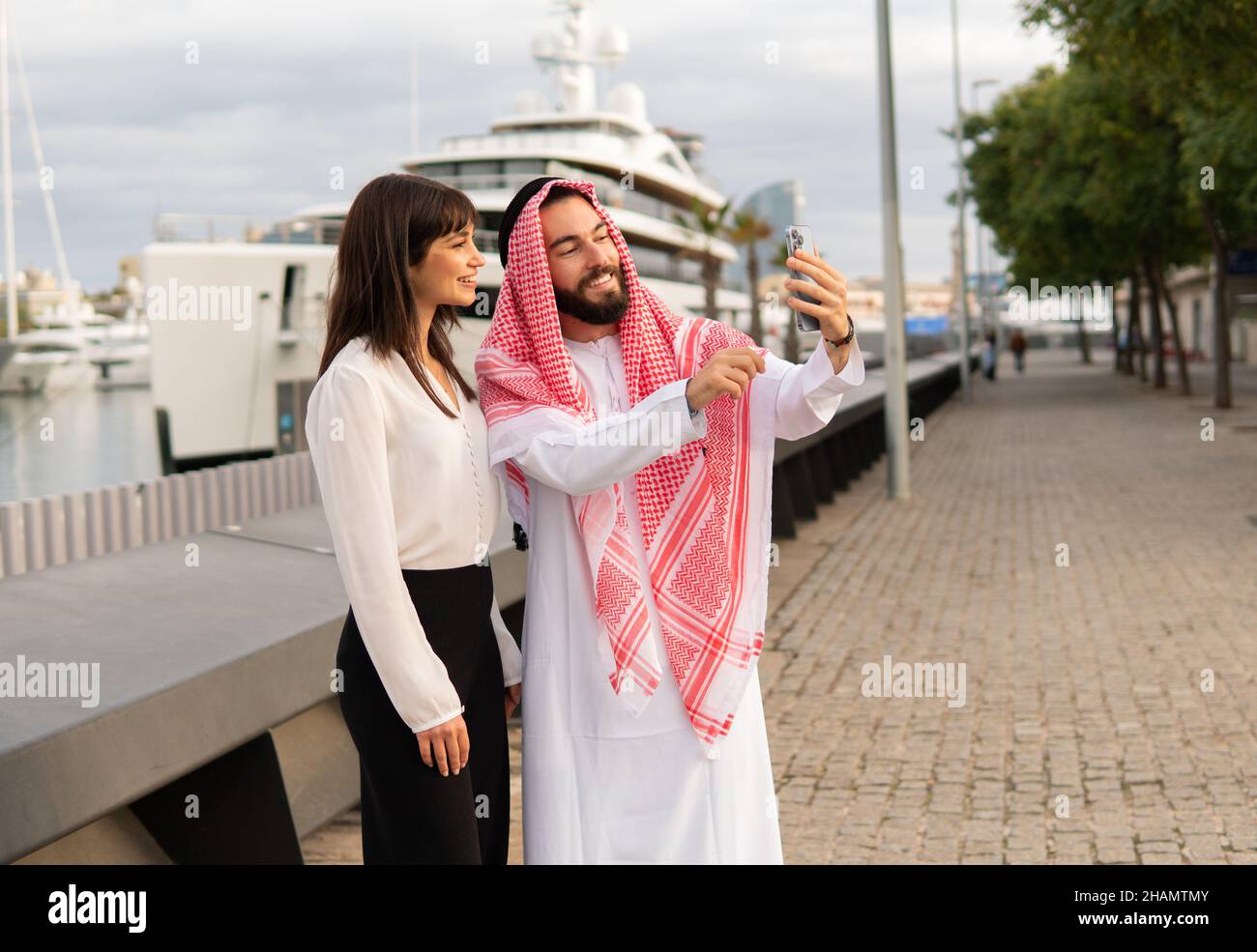 Homme d'affaires arabe souriant vêque de vêtements nationaux traditionnels et portant son selfie avec sa jeune femme assistante européenne tout en restant debout au port, divers partenaires d'affaires posent pour la photo lors de réunions Banque D'Images