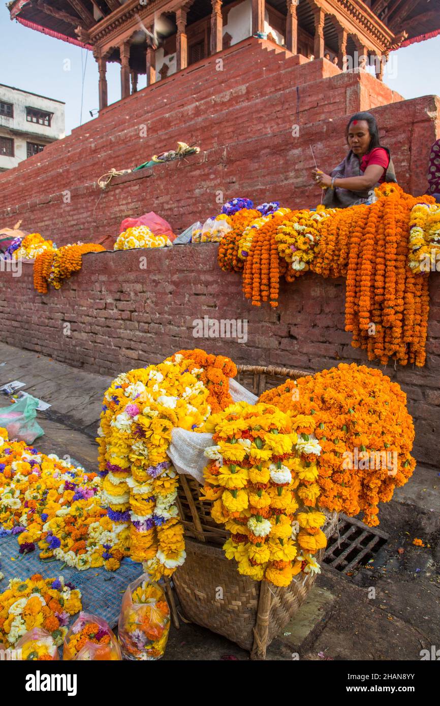 Une femme qui fait des guirlandes de fleurs pour des offrandes religieuses hindoues à vendre sur une plinthe de temple.Place Durbar à Katmandou, Népal. Banque D'Images