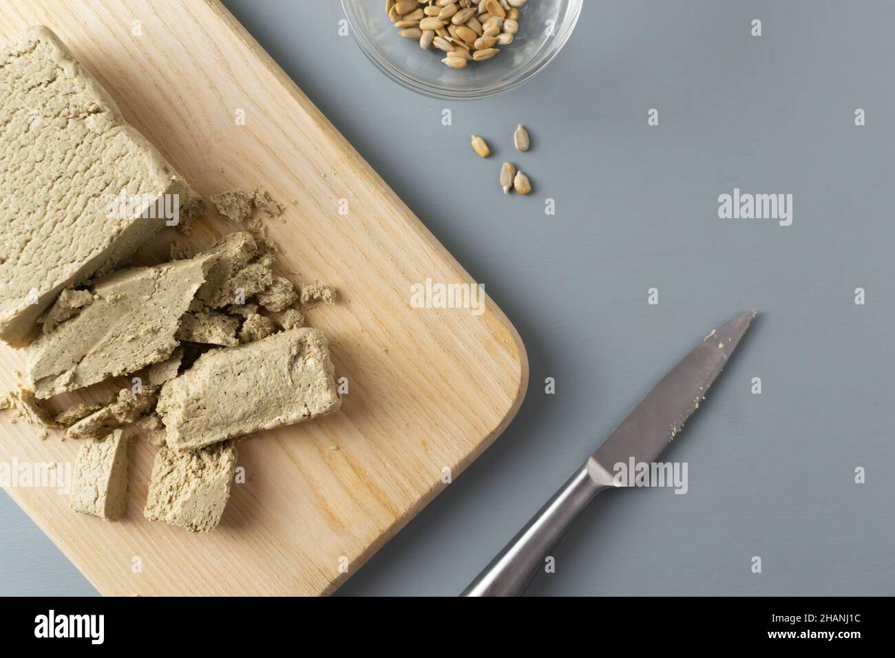 La halva de tournesol émiettée repose avec un couteau métallique sur une planche à découper en bois sur fond gris.Concept de bonbons végétariens.Orientation horizontale. Banque D'Images