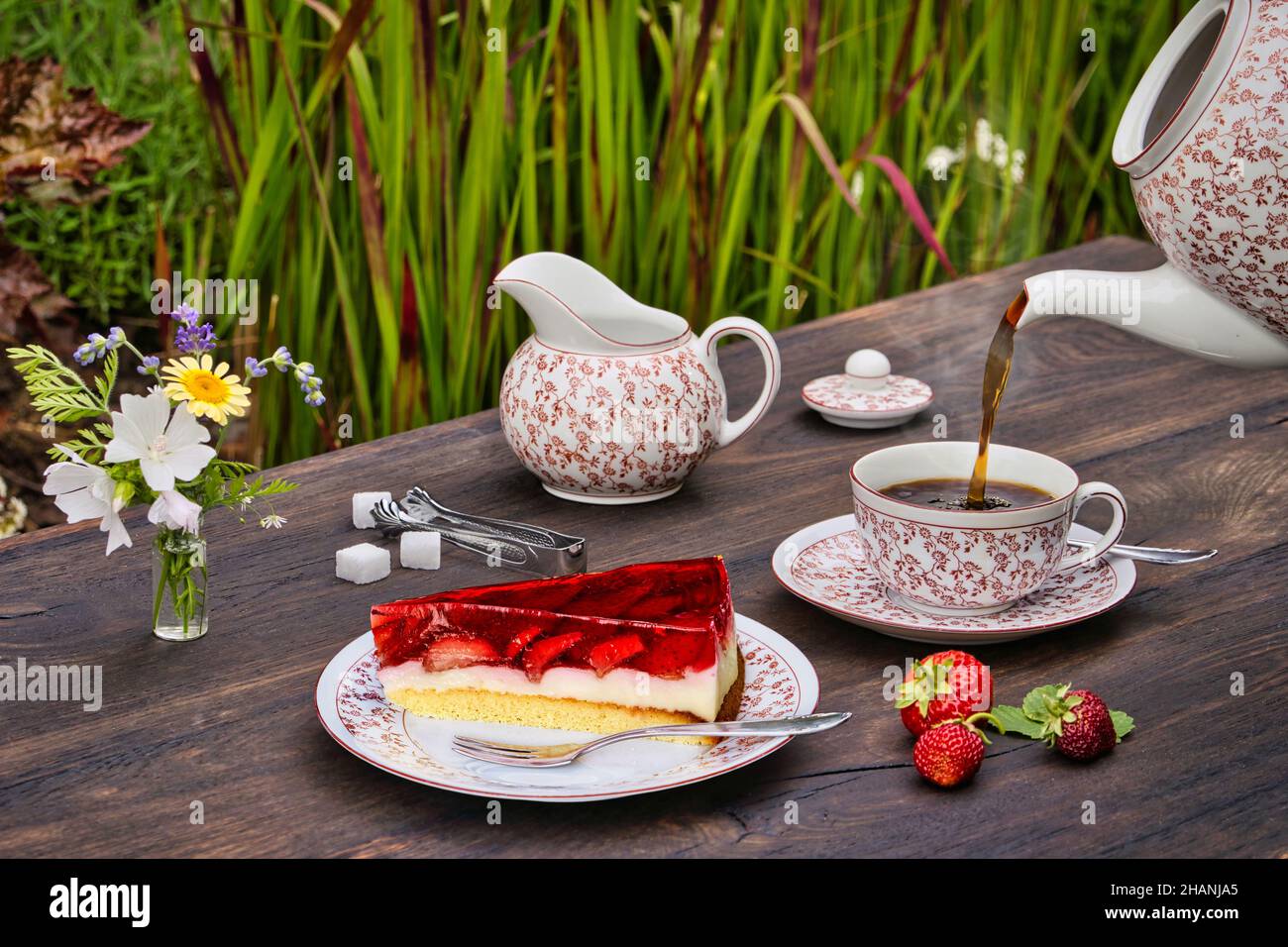 Gedeckter Kaffeetisch aus Holz im Freien mit Erdbeerkuchen, Erdbeeren und heißem Kaffee der aus einer Kaffeekanne in eine Tasse gegossen wird. Banque D'Images