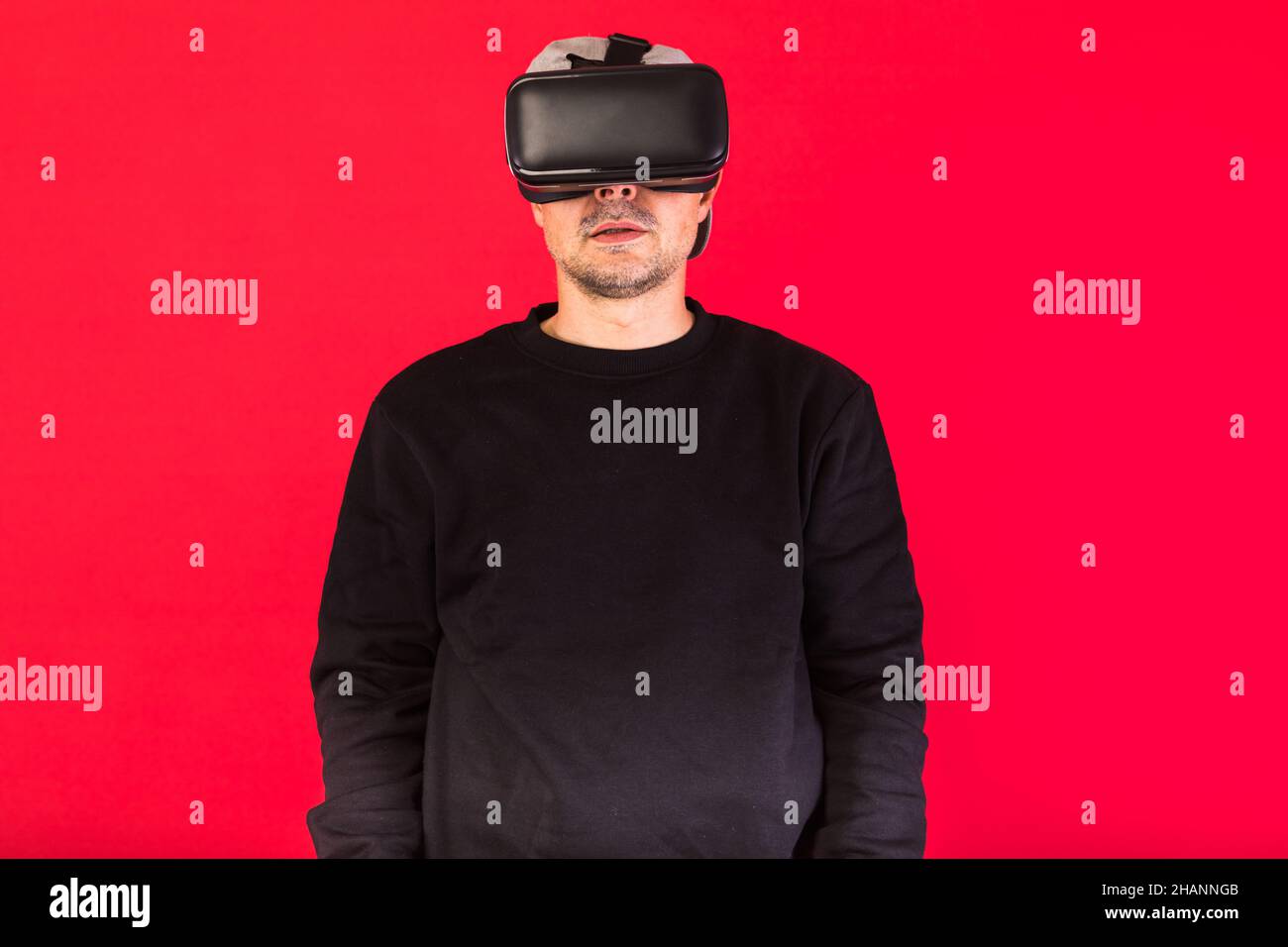 Jeune homme à barbe courte, portant un sweat-shirt noir, une casquette et des lunettes de réalité virtuelle stupéfait, regardant droit devant, sur fond rouge.Technologie, V Banque D'Images