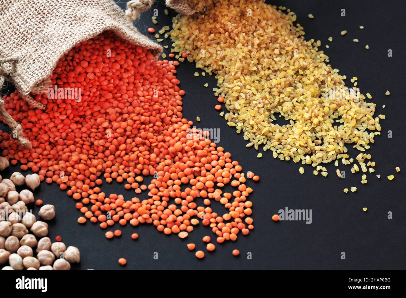 Lentilles rouges, pois chiches, riz blanc et bulgur versant du sac de jute sur la surface noire avec l'espace de copie. Concept de récolte Banque D'Images