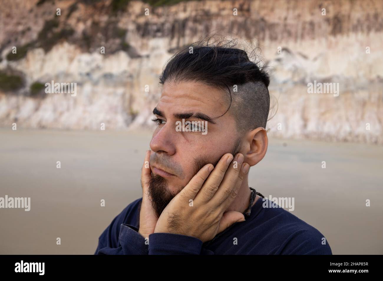 Jeune homme d'apparence hipster ou hippie avec les deux mains tenant son visage en préoccupation sur la plage Banque D'Images