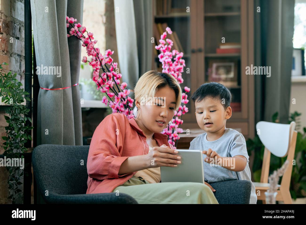 Jeune femme asiatique contemporaine montrant son mignon petit fils dessin animé en ligne tout en ayant le repos à la maison Banque D'Images