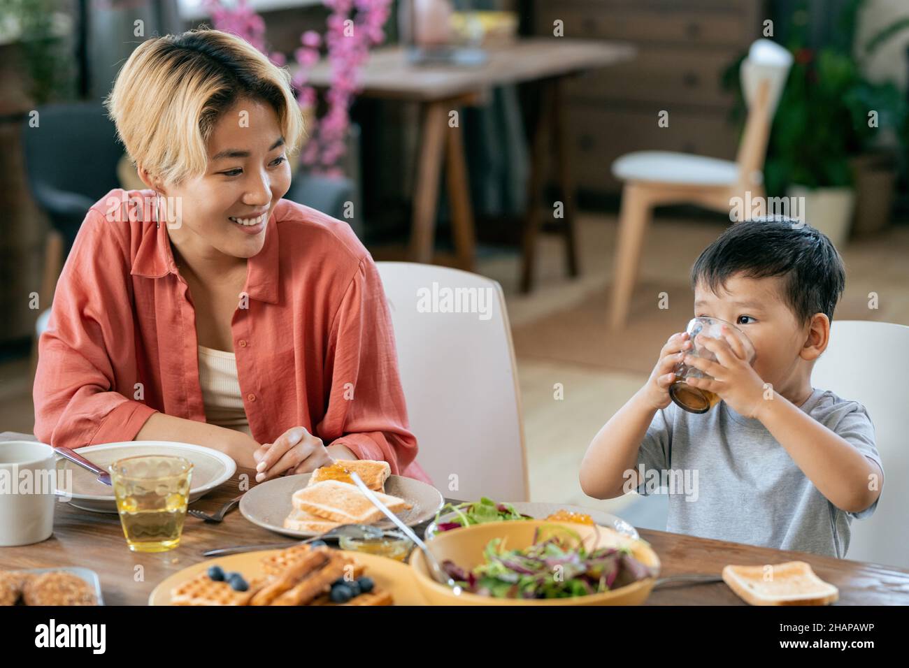 Bonne jeune femme asiatique regardant son adorable petit fils boire du thé ou du jus au petit déjeuner Banque D'Images