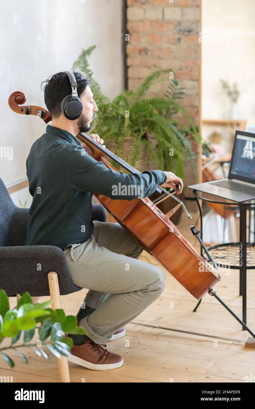 Jeune professeur de musique jouant du violoncelle devant un ordinateur portable pendant une leçon à la maison ou une formation pour le public en ligne Banque D'Images